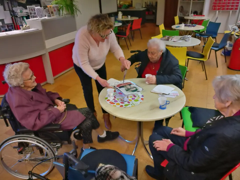 Ældre medlemmer, en af dem i kørestol, samlet om et cafébord med farverige stole
